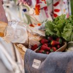 A woman in a floral dress holds a basket filled with bread, strawberries, and lettuce on her bicycle.