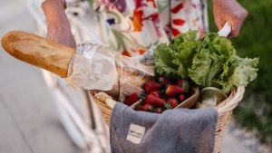 A woman in a floral dress holds a basket filled with bread, strawberries, and lettuce on her bicycle.