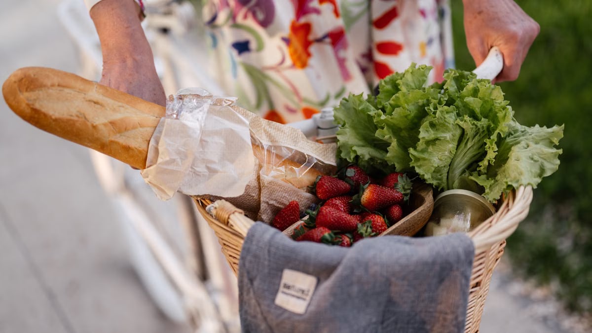 A woman in a floral dress holds a basket filled with bread, strawberries, and lettuce on her bicycle.